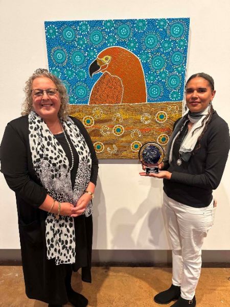 Two people pose in an art gallery in front of a colorful painting of a large bird. One person holds a blue glass award as they smile for a commemorative photo celebrating an achievement.
