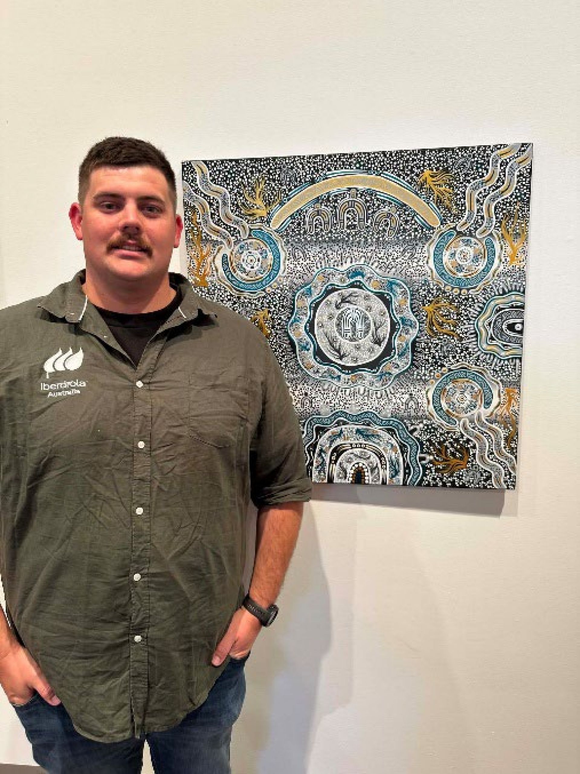 A smiling person poses in an art gallery beside a detailed Aboriginal painting featuring circular designs, flowing waterways, and intricate dot work in blue, white, gold, and black.