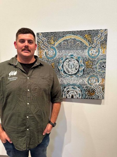 A smiling person poses in an art gallery beside a detailed Aboriginal painting featuring circular designs, flowing waterways, and intricate dot work in blue, white, gold, and black.