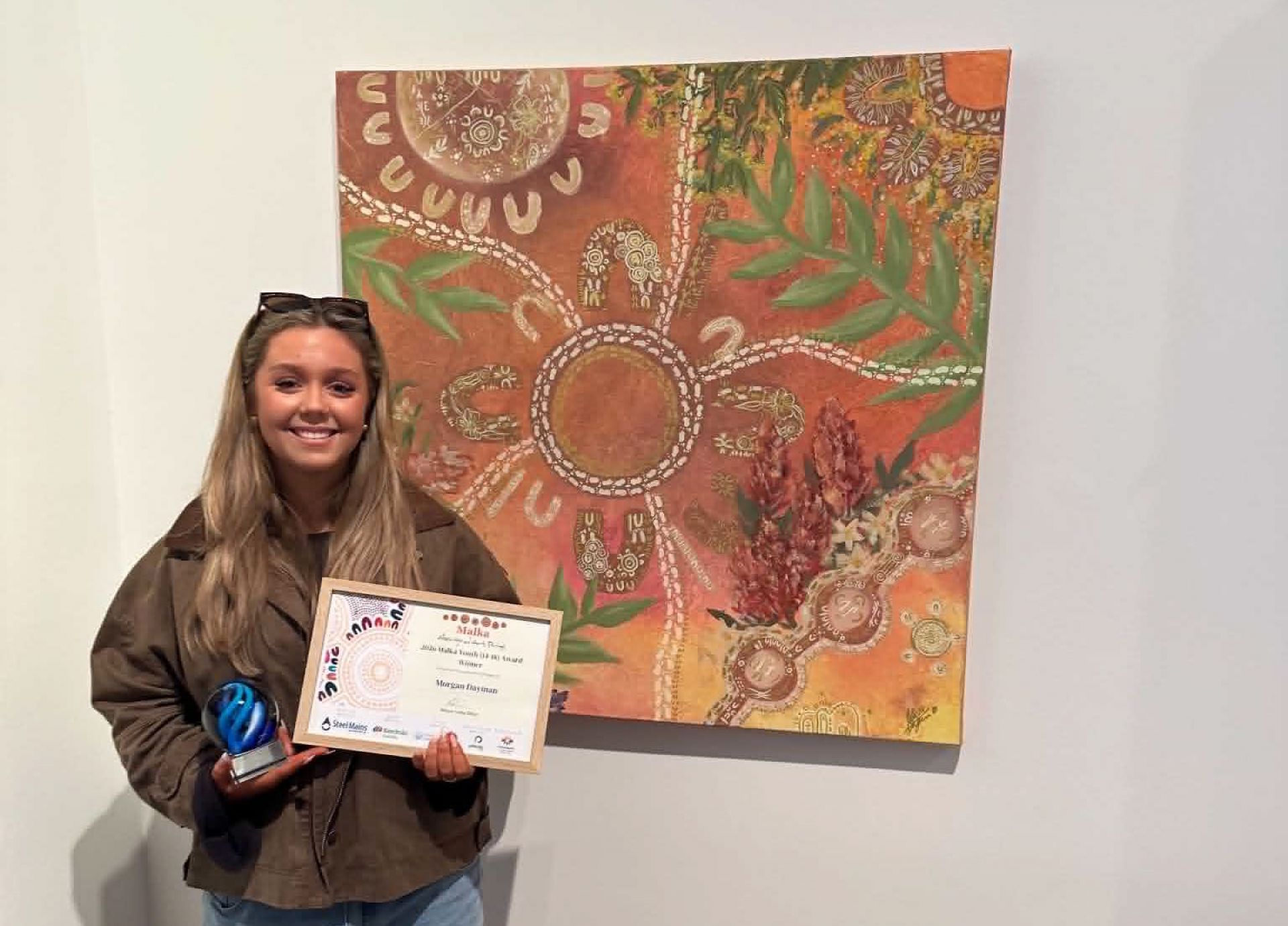A smiling award recipient stands beside a large Indigenous-inspired painting in an art gallery, holding a blue glass trophy and a framed certificate while posing for a celebratory photo.