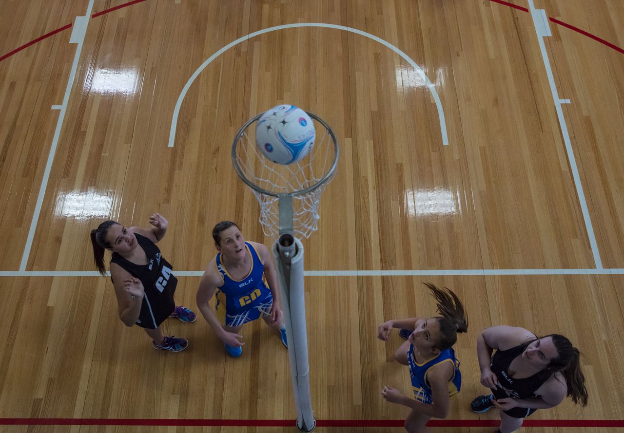 Photograph from above of four netball players watching a netball pass through the goal hoop. Two of the players are in black and white uniforms and two of the players are in blue and gold uniforms.